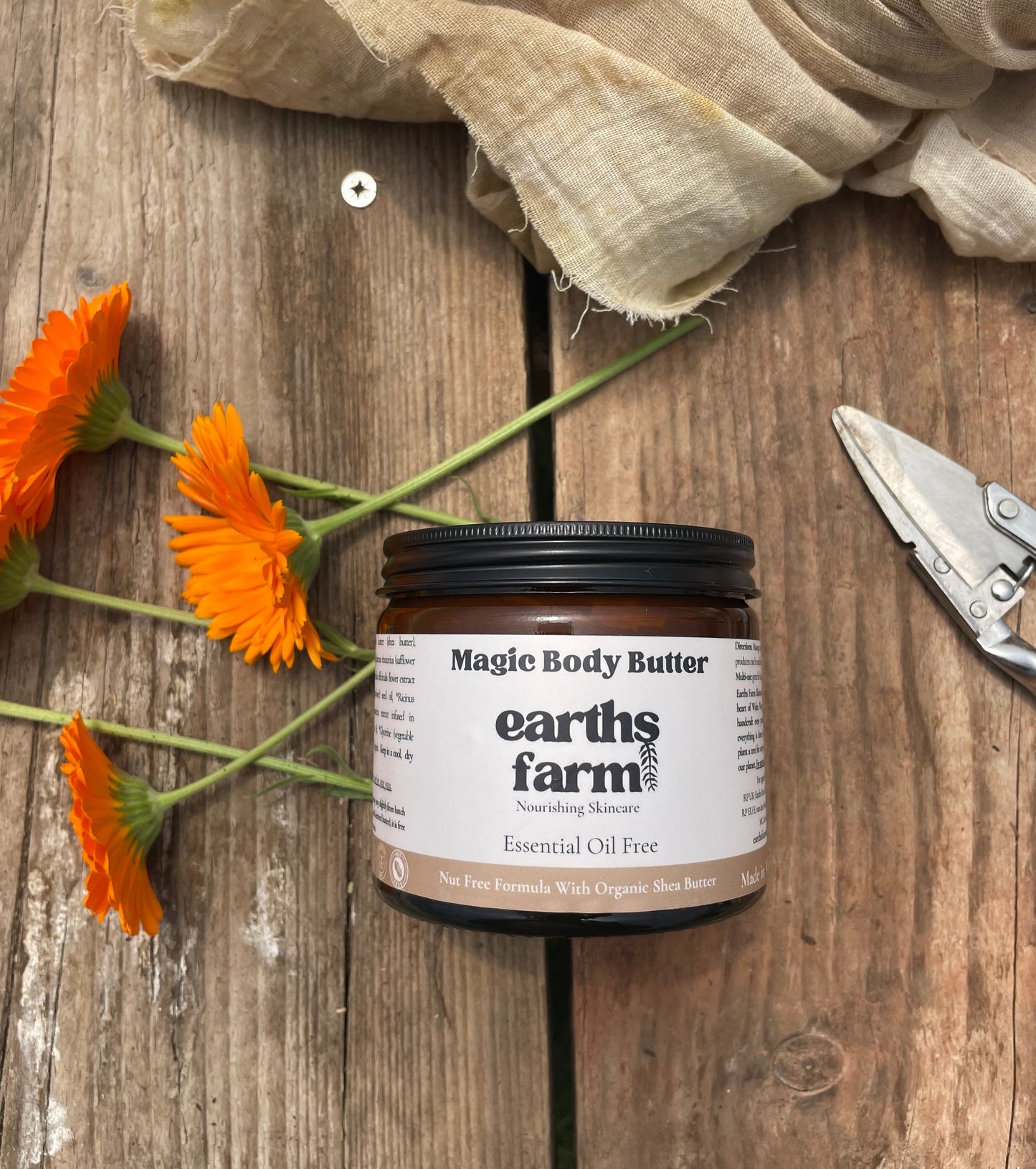 Jar of Magic Body Butter from Earth's Farm on a wooden surface with orange flowers and a cloth.