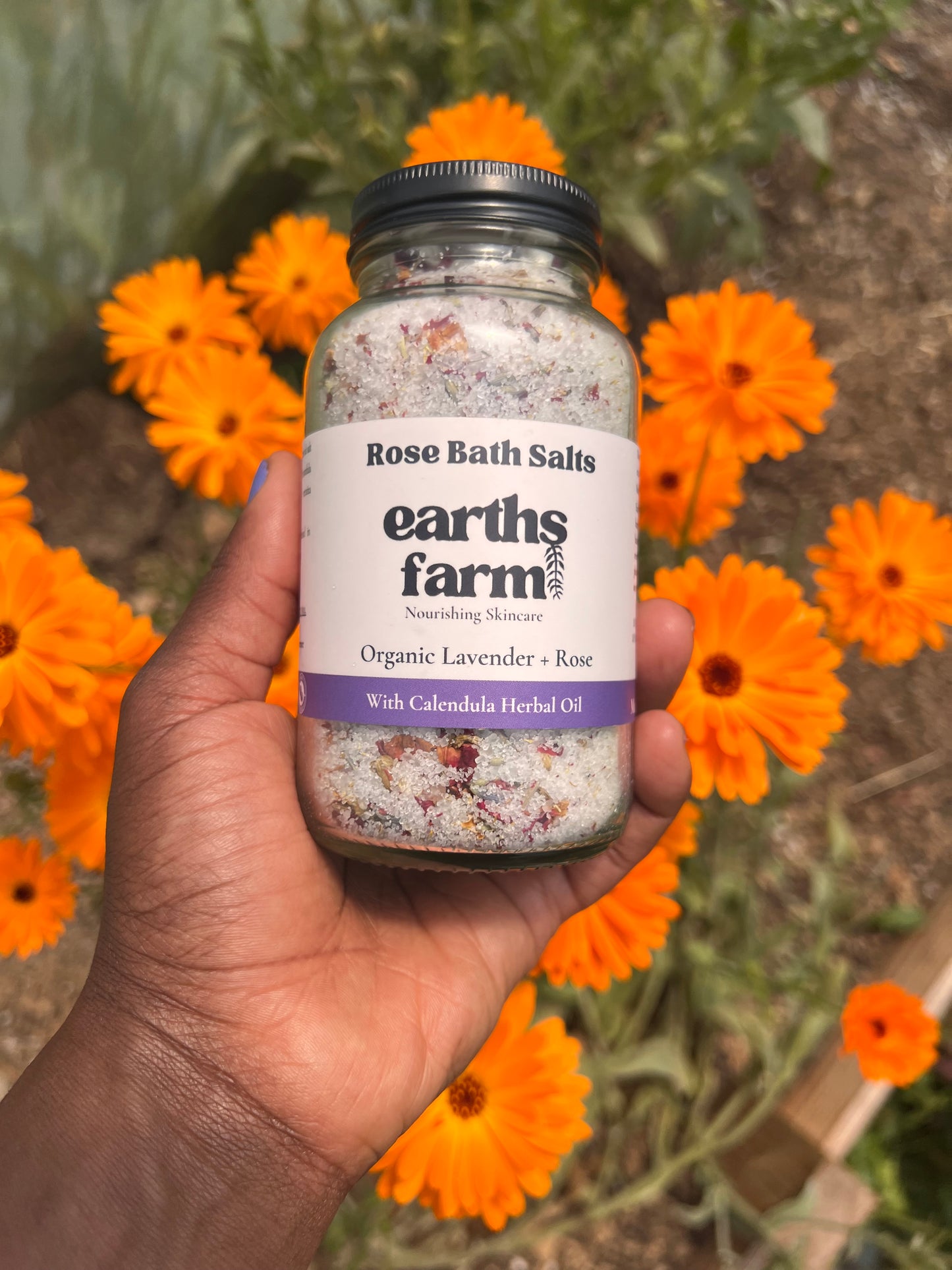 Hand holding a jar of 'Earths Farm' rose bath salts with orange flowers in the background