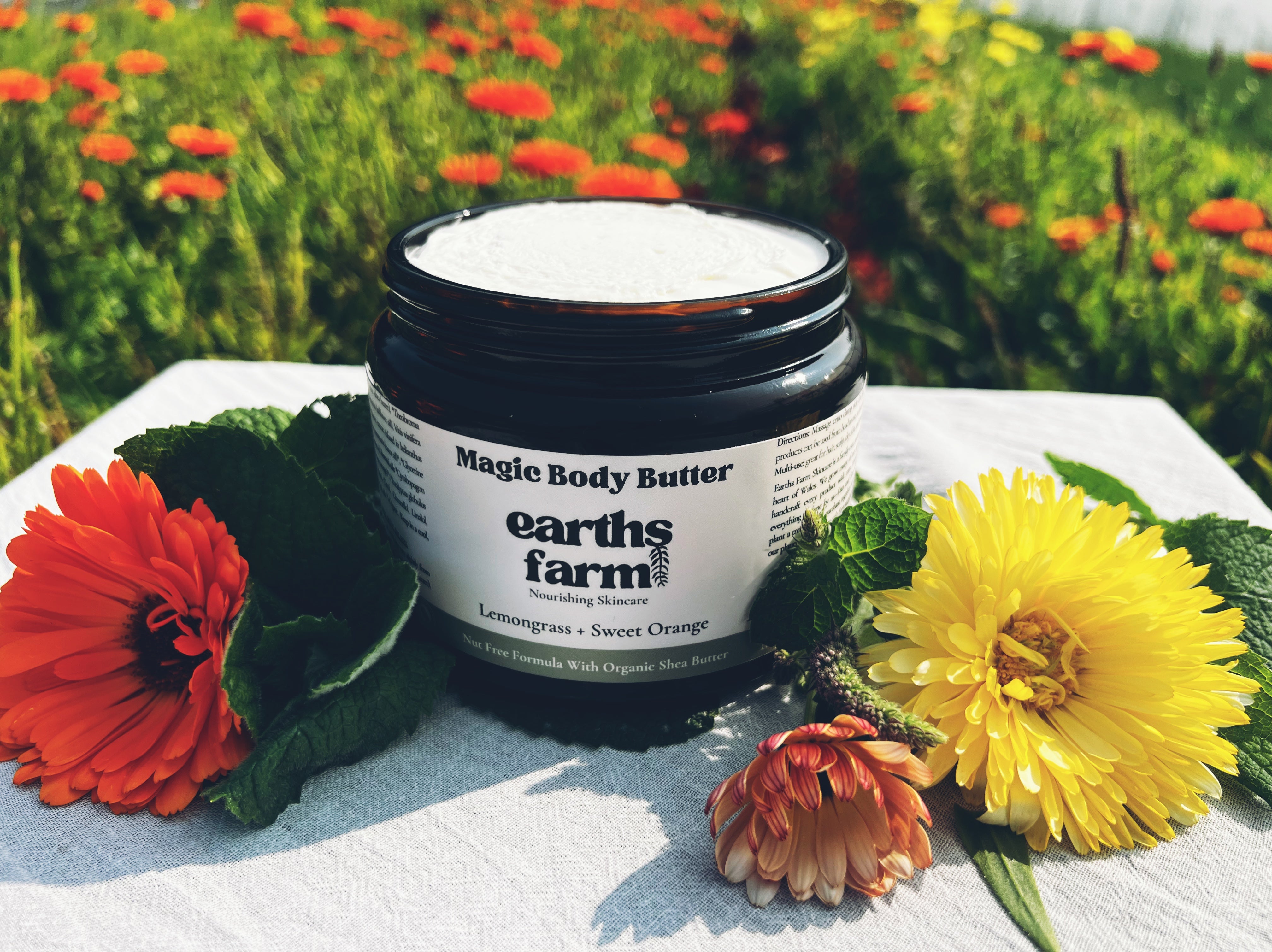 Jar of 'Magic Body Butter' from 'earths farm skincare surrounded by flowers on a white surface with a natural background.