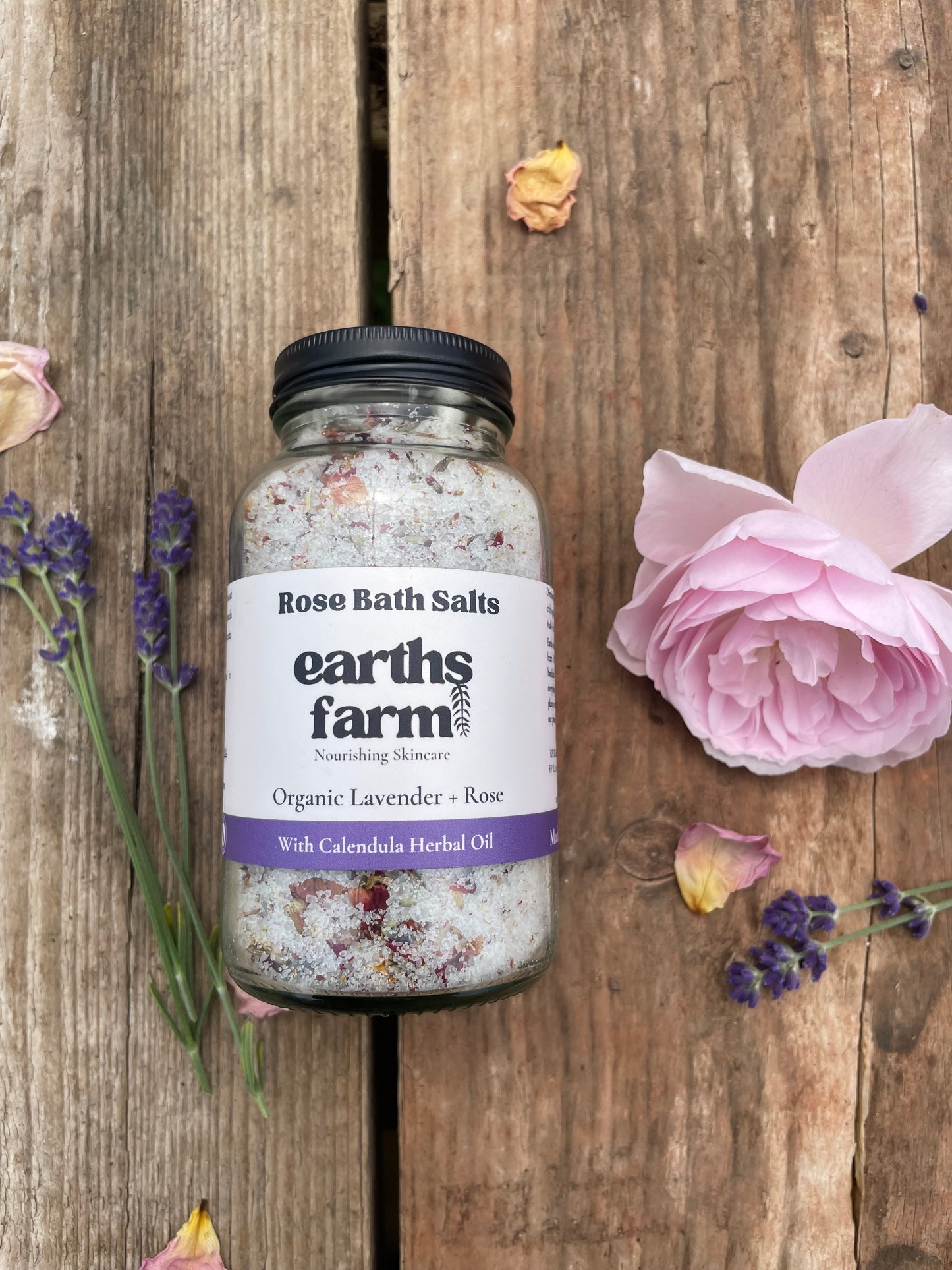 Bottle of Earth's Farm rose bath salts on a wooden surface with flowers.