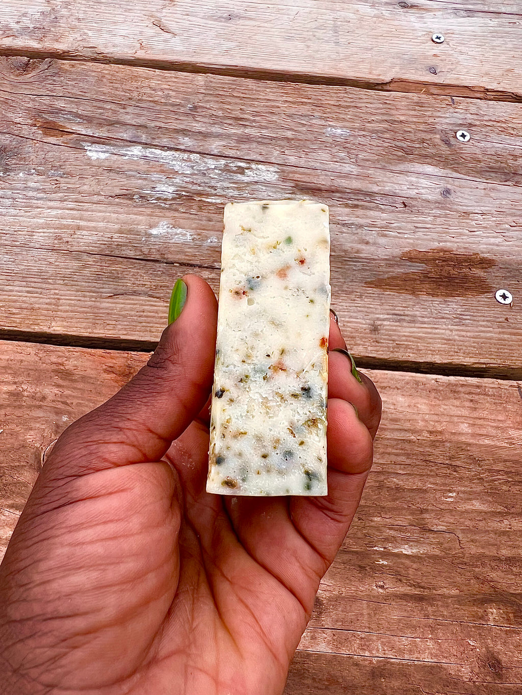 Hand holding a rectangular block of soap with visible herbs on a wooden surface
