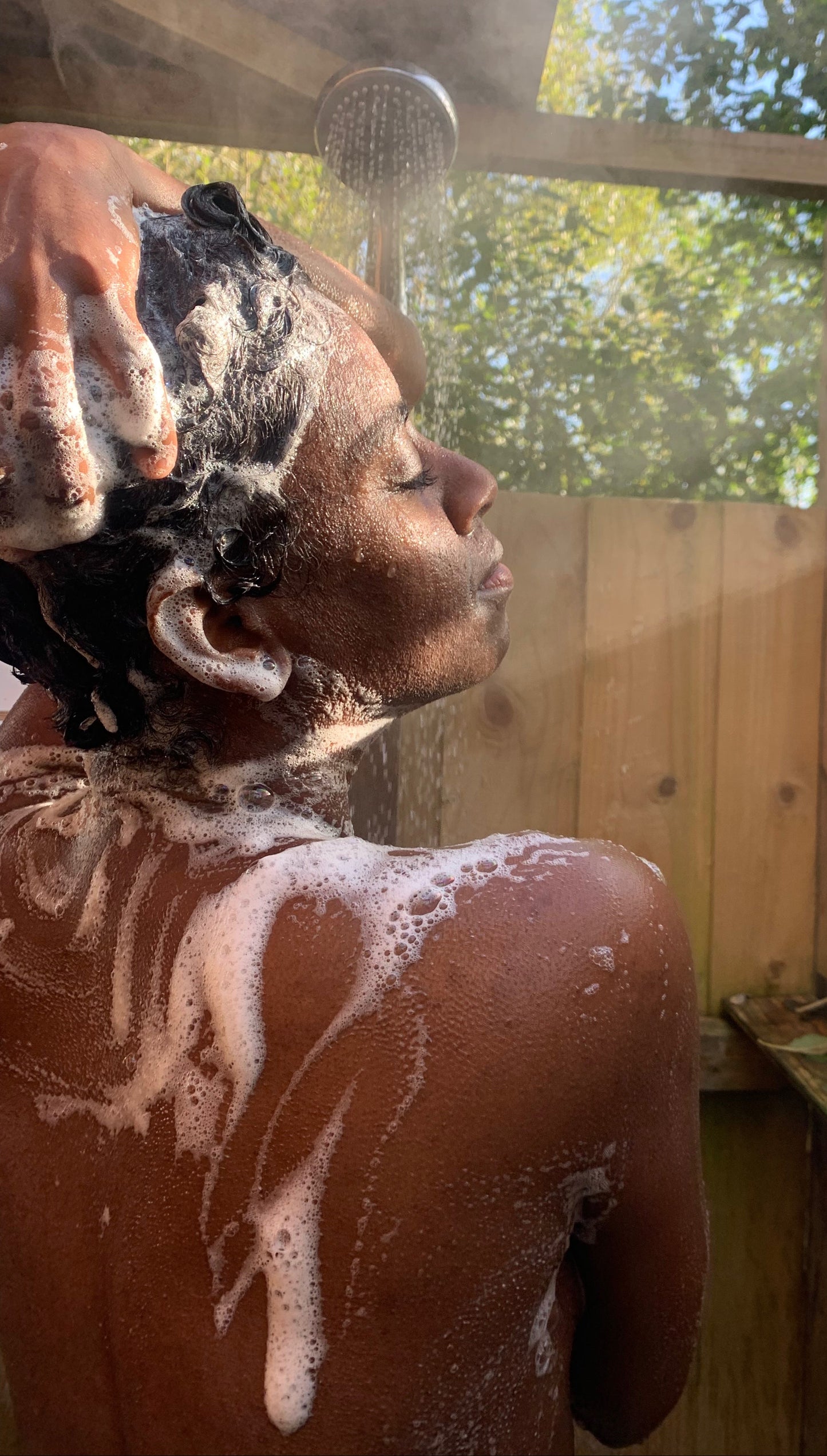 Person washing hair outdoors with a wooden shower enclosure and trees in the background